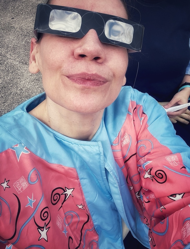 a womann in a pink and blue jacket looking up with eclipse glasses on