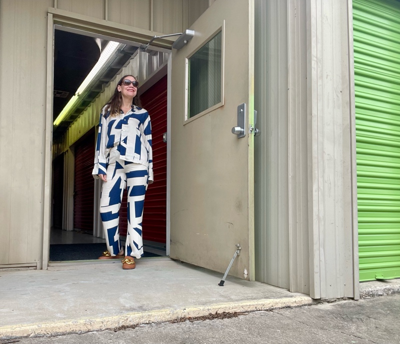 a woman in a blue and white linen pant and white long sleeve pant set in front of green doors and concrete walls  