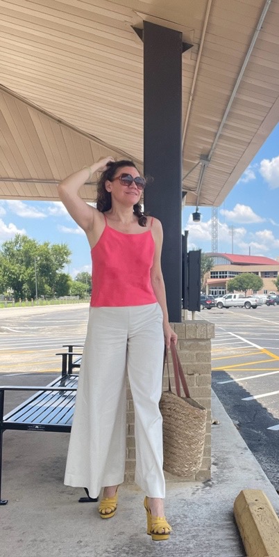 a woman in a coral tank top and white pants and yellow wedges with a straw bag