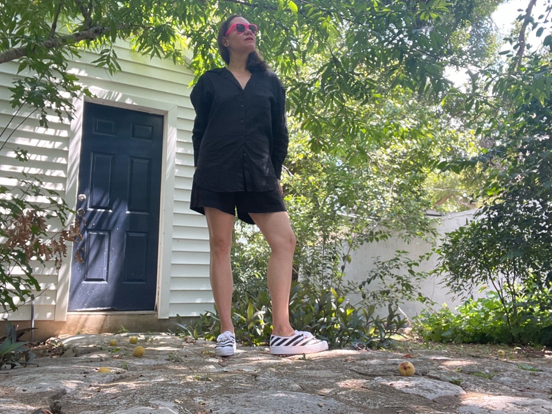 a woman in black linen shorts and a button down with black and white sneakers and pink sunglasses